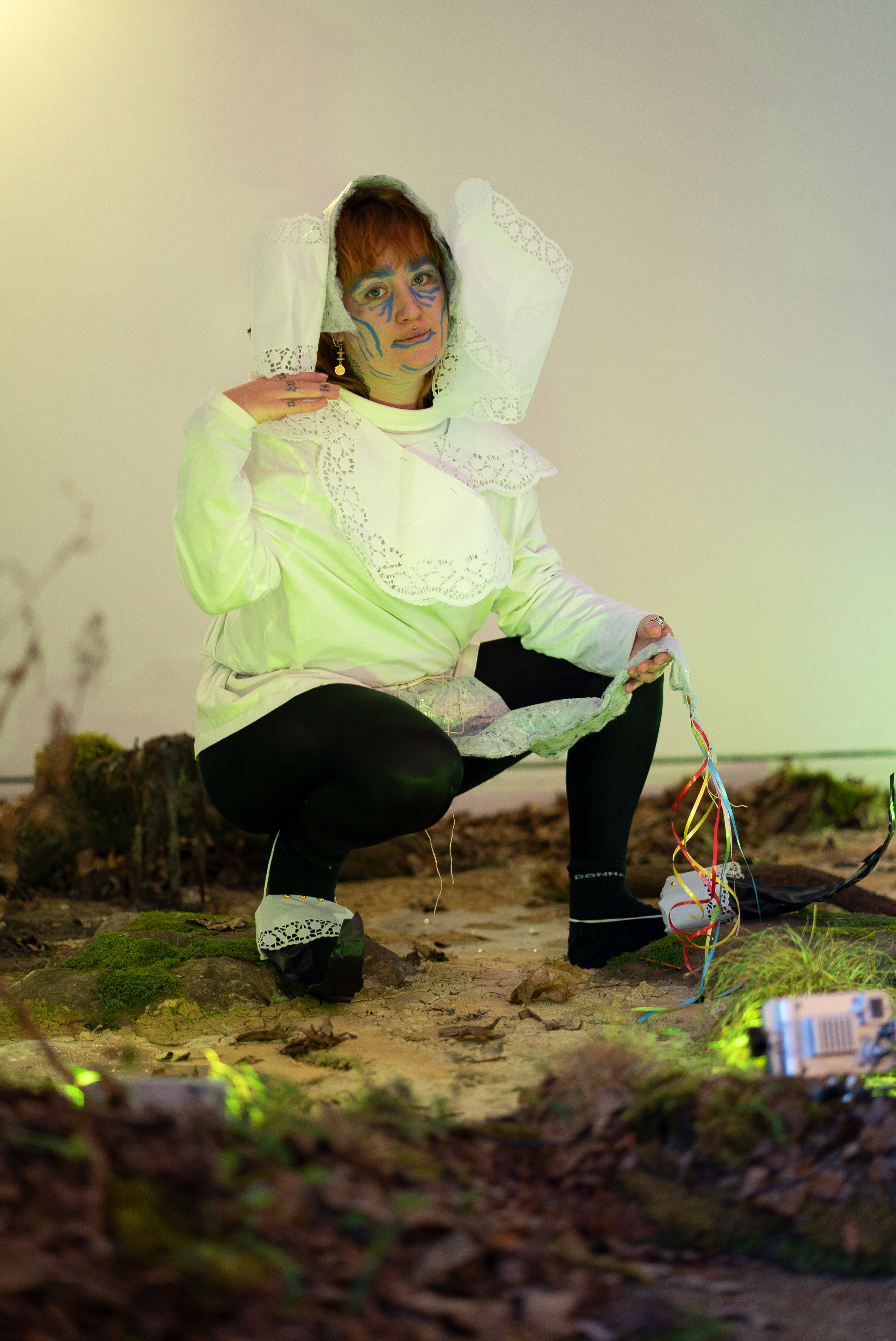 The photo shows a person posing in a squatting position. They are wearing a headdress and a top made of white lace, black leggings and homemade goblin shoes made of black gaffer and white lace. They have put their right hand on the right shoulder and look directly into the camera. The person's body is illuminated by a green light on the ground. The ground is an artificial swamp, which consists of soil, moss, dried leaves and grass. The background is a white wall.