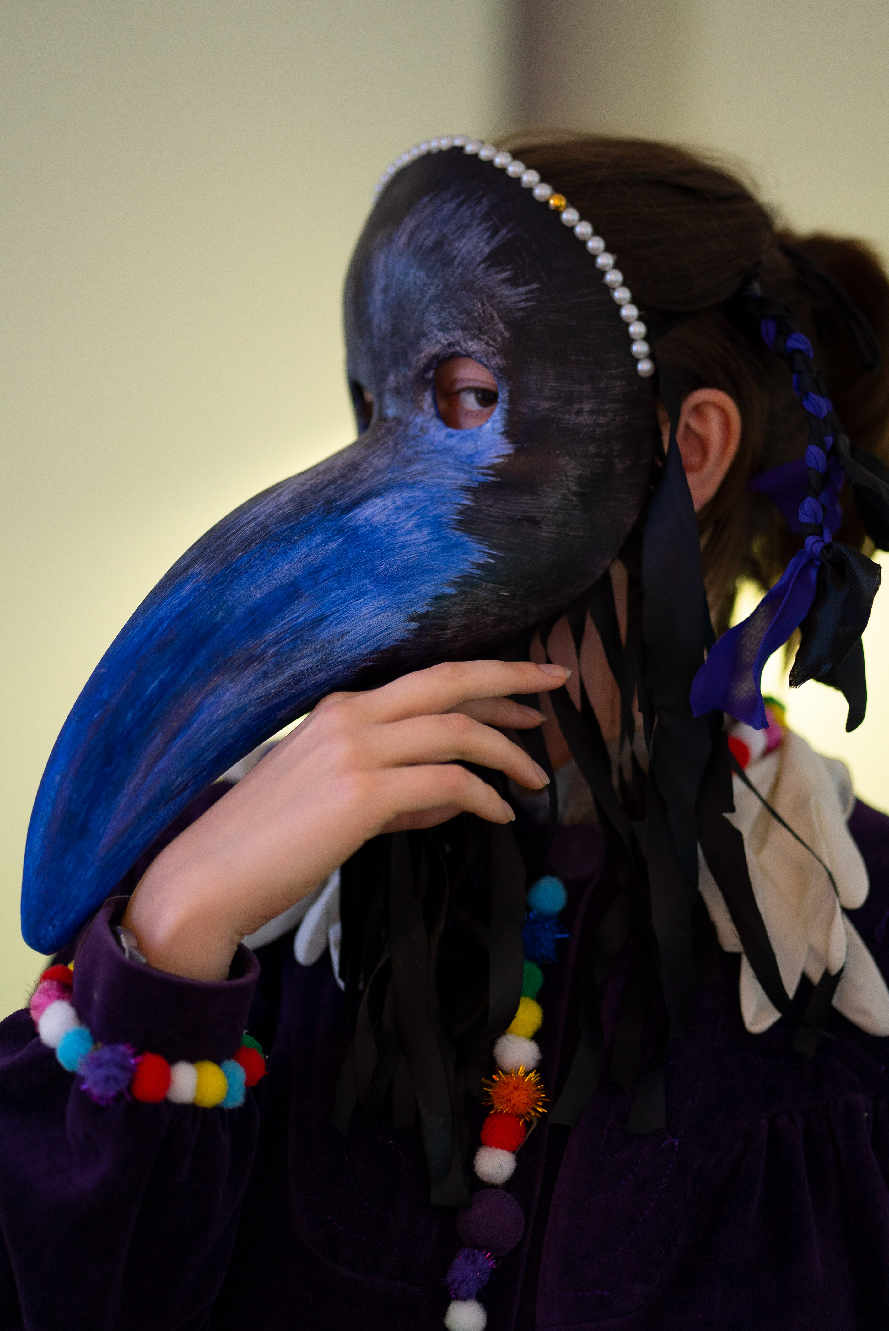 The photo is a close-up of a side profile of a person wearing a plague doctor mask. The mask is painted with black and blue paint, the upper edge of the mask is decorated with white pearls. The person is wearing a purple velvet coat decorated with colorful fabric balls on the cuffs and button closures. They hold their right hand under the beak of the mask. The eye visible under the mask looks directly into the camera.