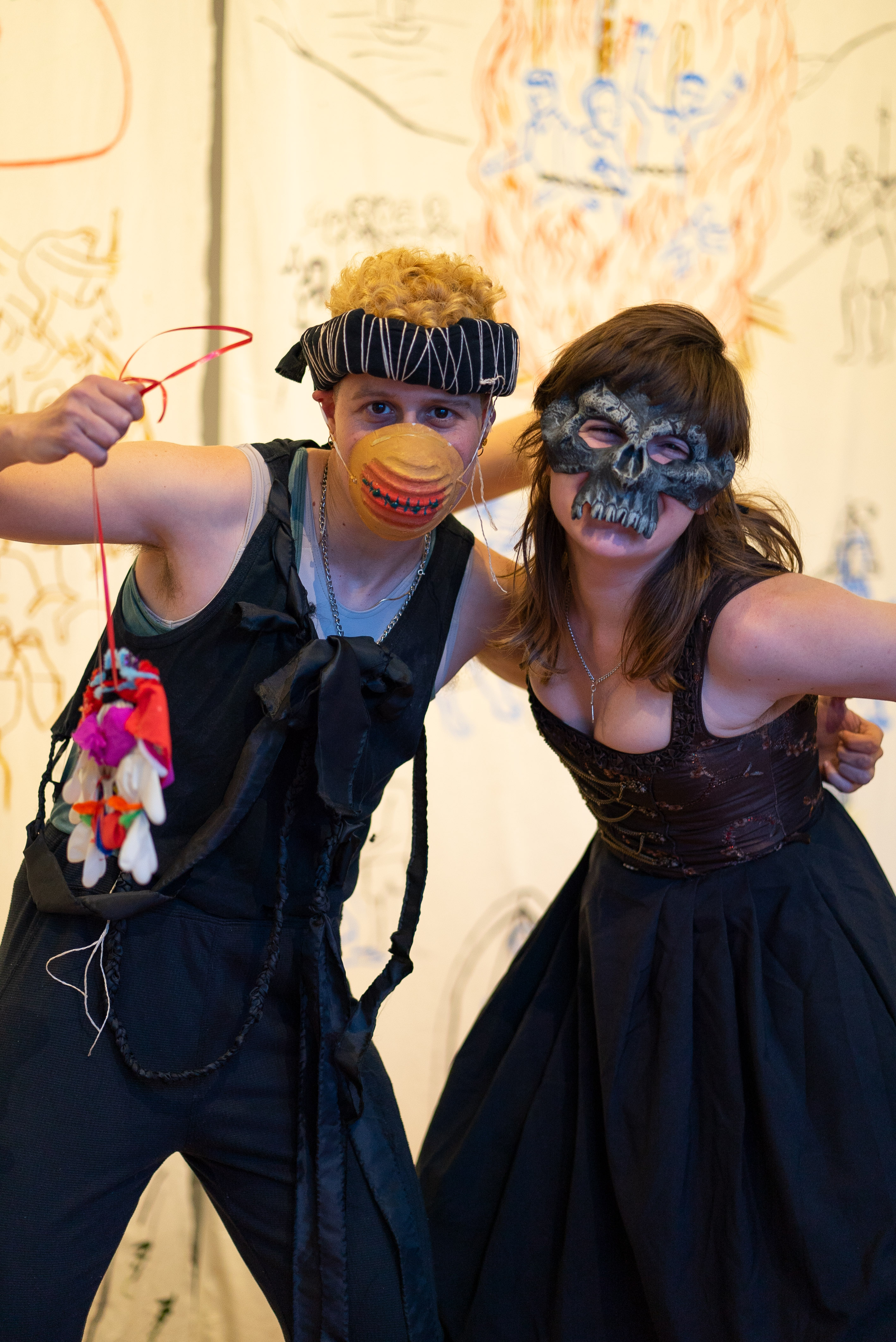 The photo shows two people in costumes posing on a stage. They have one arm around each other. The person on the left wears a patterned fabric ring as headgear, a painted protective mask that looks like a sewn up mouth, a black muscle shirt and black trousers. In their right hand they hold a self-made medallion in the air. The right person is wearing a skull mask and a dress that accentuates their cleavage. In the background hang banners made of antique white fabric with colourful medieval figurative paintings.