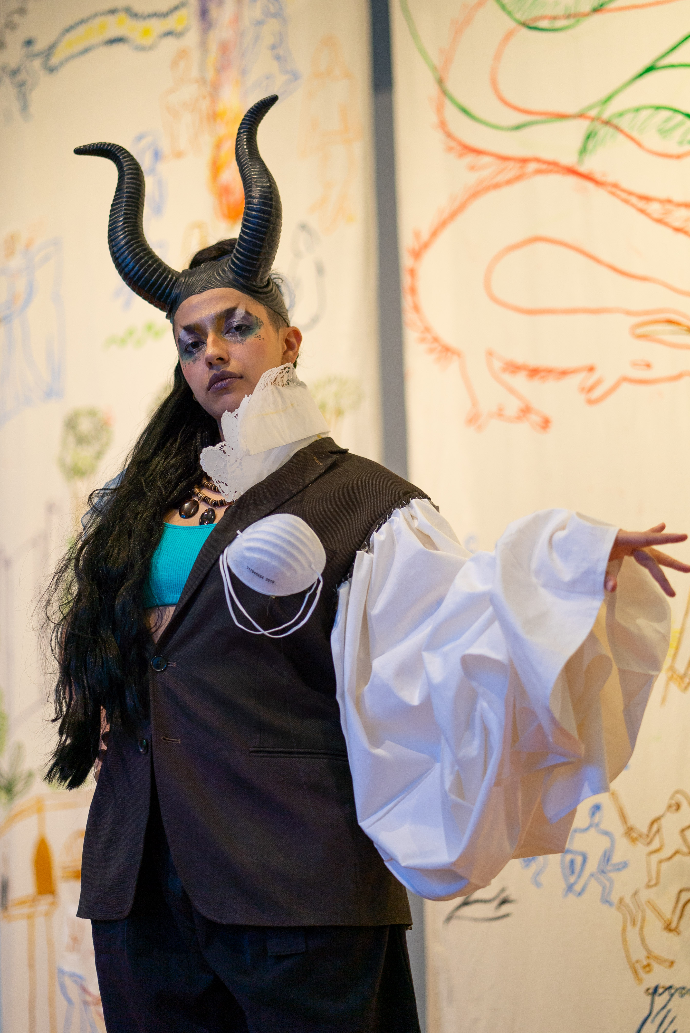 The photo shows a person posing in an imaginative costume. They wear two plastic horns on her head and a long black plait of hair down to her hips. Their left hand is outstretched to the side and shows a voluminous white sleeve. In the background we see antique looking white banners painted with colored figures by the artist Irene Fernandez Arcas.