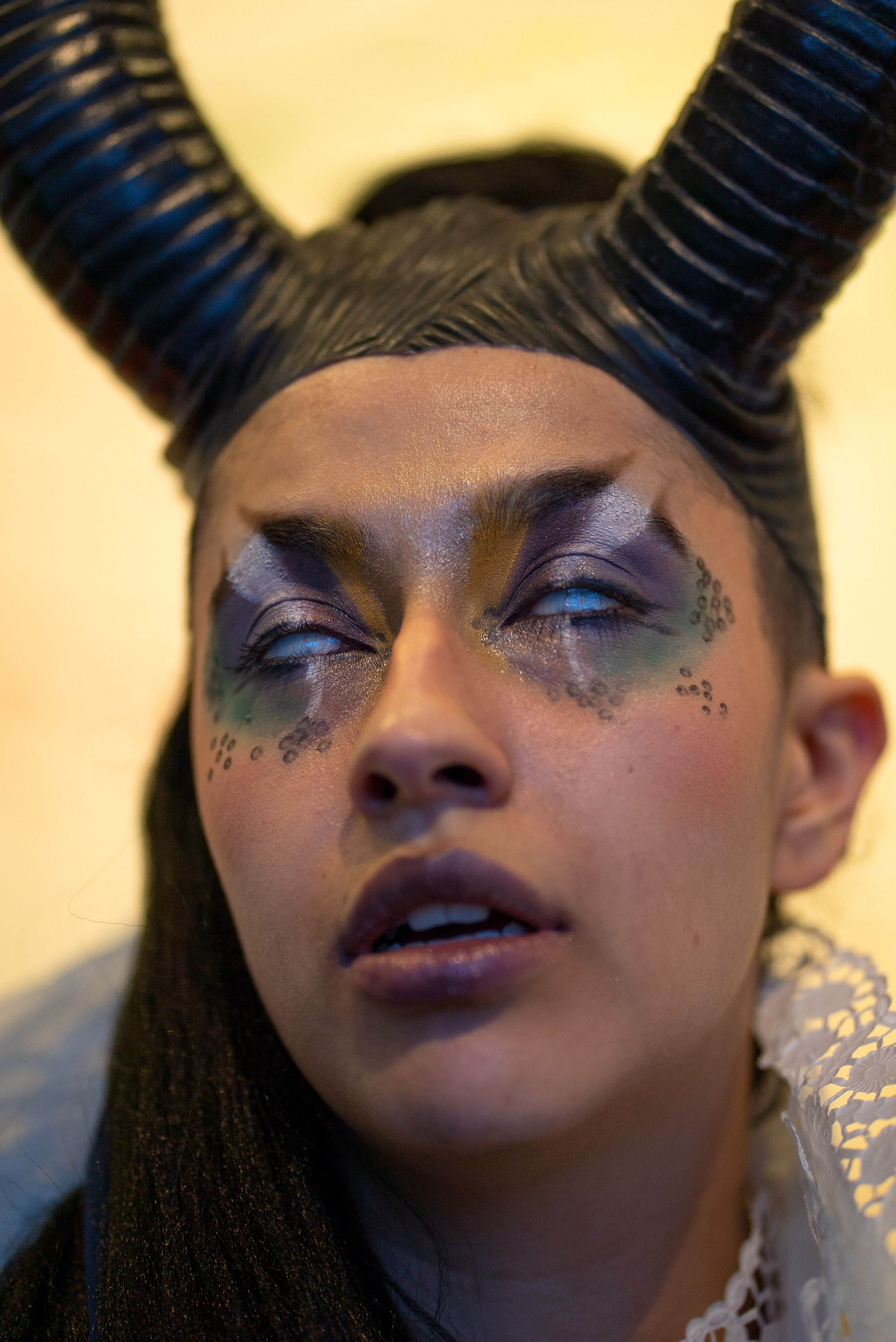 The photo is a close-up of a person with two plastic horns as headgear and imaginative eye make-up in purple, green, white and black with glitter. They have opened their mouth slightly. Their eyeballs are turned upwards and slightly opened, so that only the white of the eye is visible.