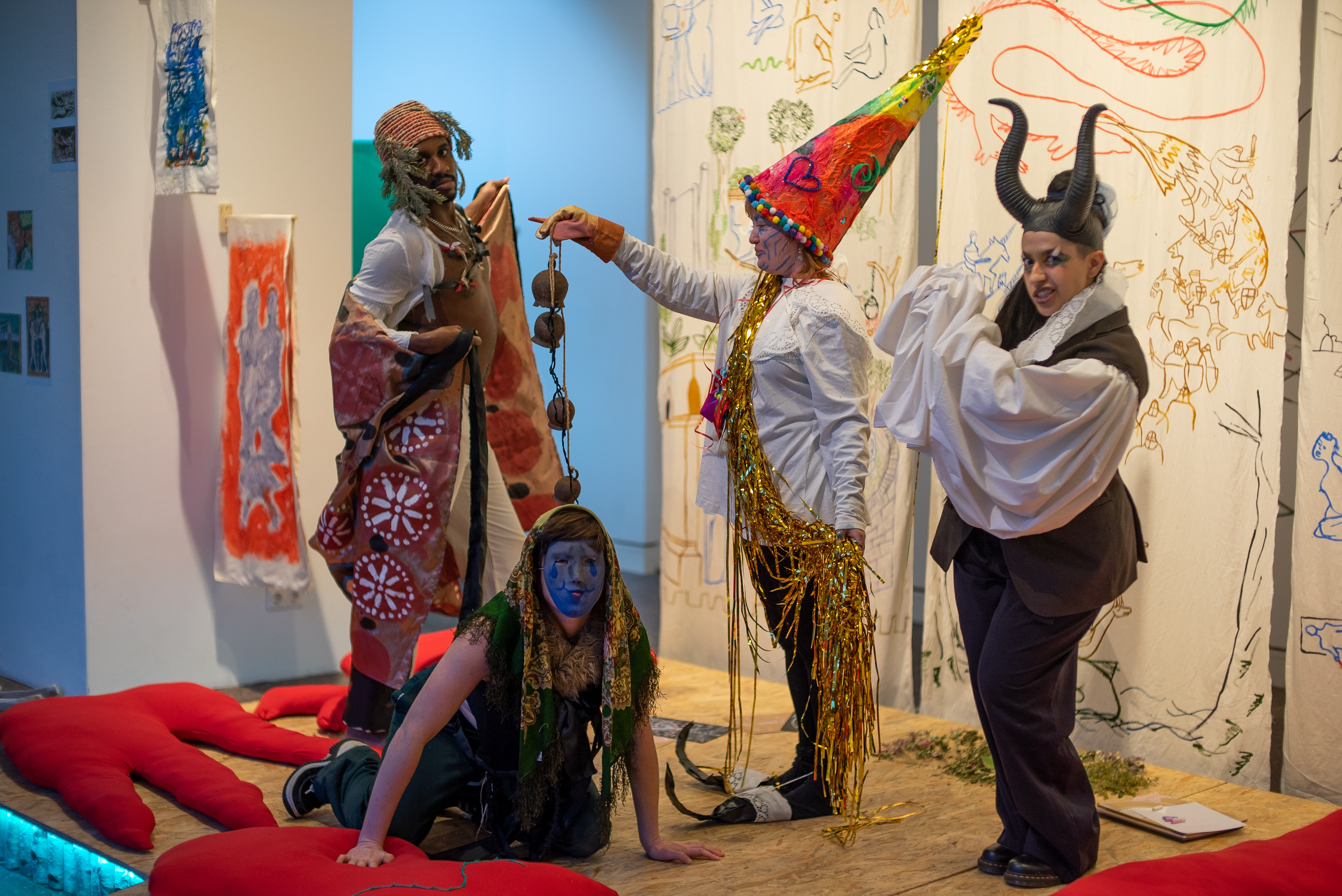The photo shows four people on a stage in imaginative medieval costumes. Three persons pose standing, the fourth kneels on the floor in front. In the background we see three banners made of antique white fabric, which were painted by the artist Irene Fernandez Arcas with figures made of brushstrokes. On the floor of the stage are several large red cushions in figurative shapes.