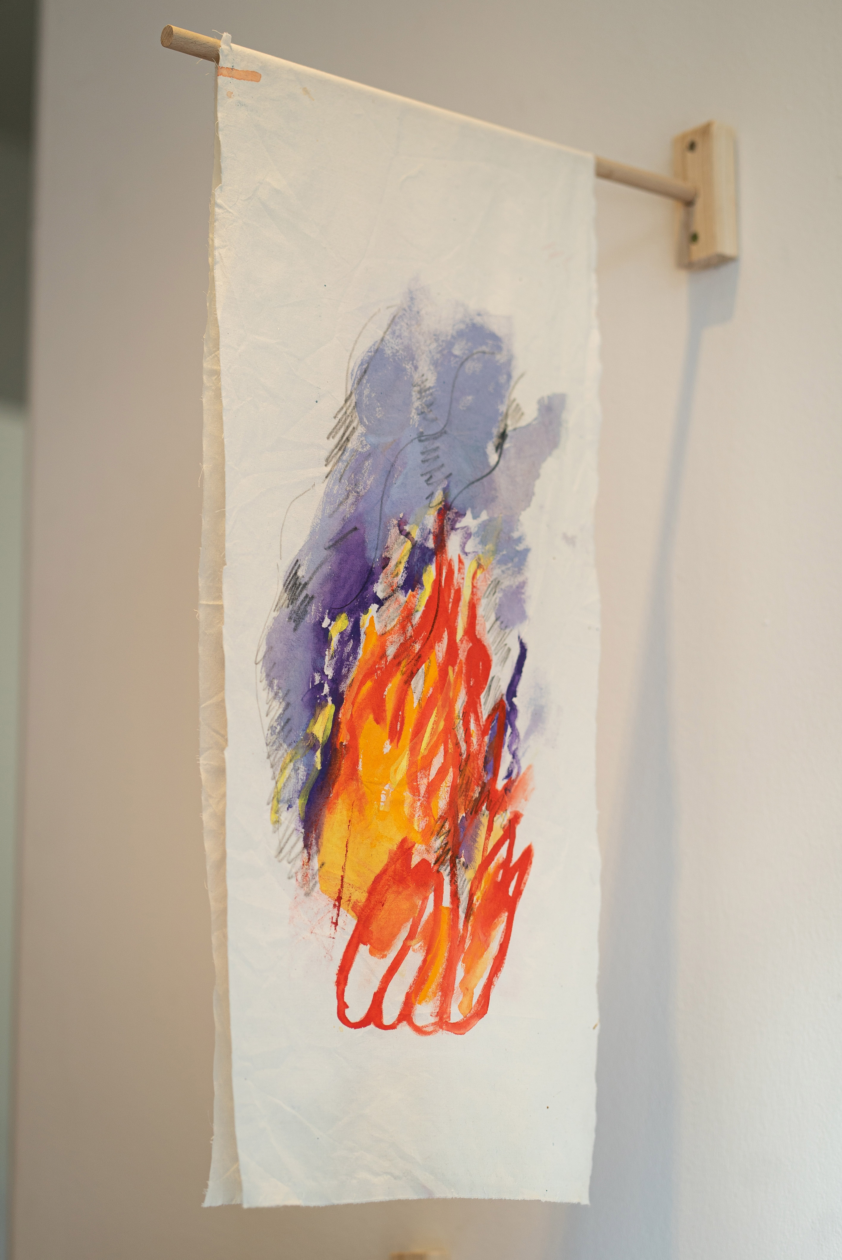 The photo shows a piece of rectangular antique white fabric hanging from a wooden pole on the wall. It resembles a coat of arms. A fire is painted on the fabric with bright red, orange and purple colors. The coat of arms is part of the stage installation by Irene Fernandez Arcas for the exhibition "Burlungis" at Galerie im Turm in Berlin.