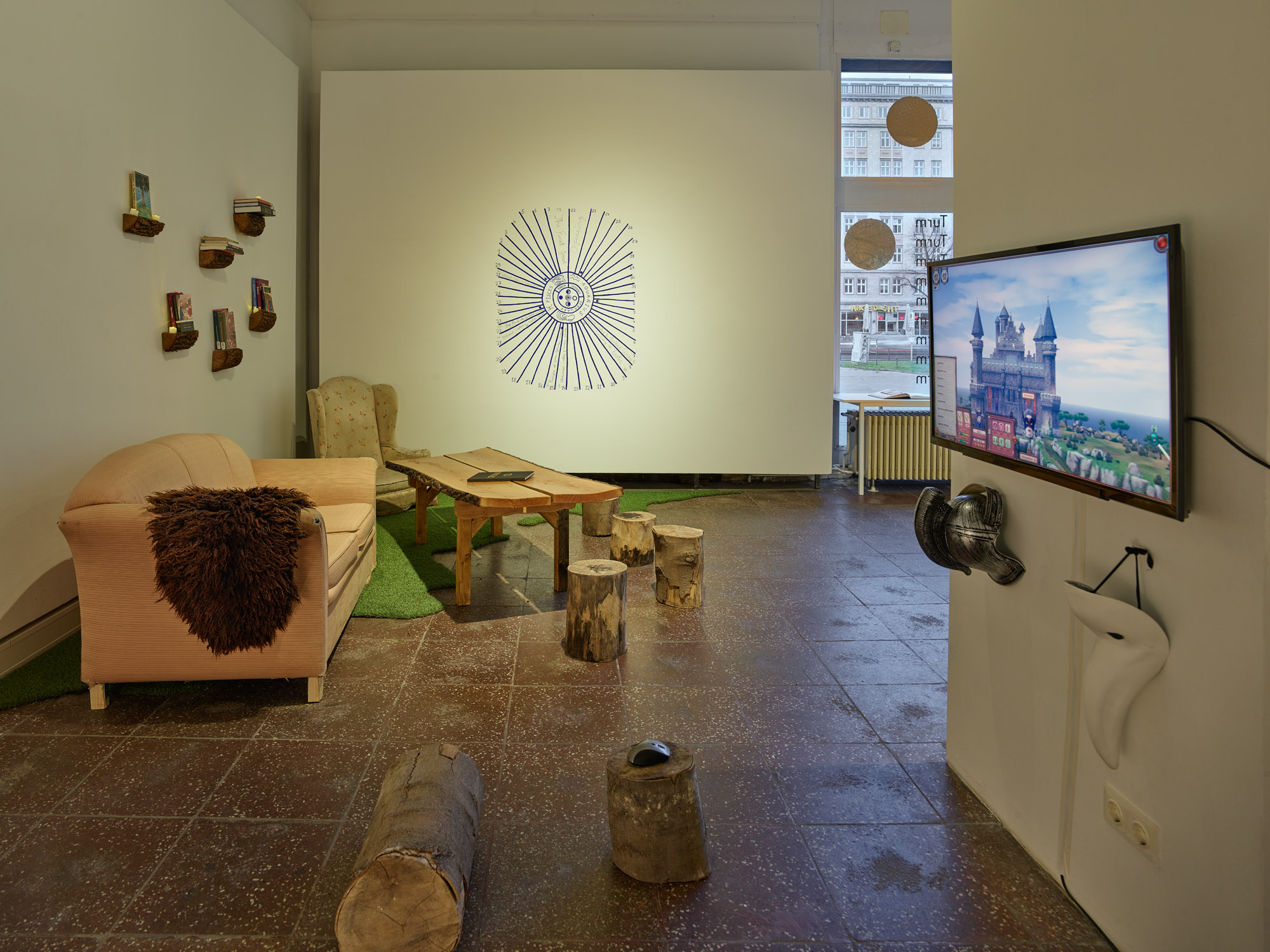 The photo is an exhibition view of the show "Burlungis" in Galerie im Turm in Berlin. On the left side of the space next to the wall there is an old pink couch, behind it in the corner a beige armchair, next to the couch a table whose table top was made from a tree trunk with bark. The furniture stands on a piece of artificial grass in a curvy shape. The rest of the floor is made of stone-gray tiles. Cut tree trunks are scattered around the table as seating. In the middle on the back wall is painted a blue color and an oval shape a lunar calendar. On the right side of the picture there is a monitor on the wall of a column. A lock can be seen on the monitor in 3D. The image section belongs to a computer game. Two objects decoratively hang under the monitor. The left object is a silver armor made of plastic, the right object is a white plague doctor mask.
