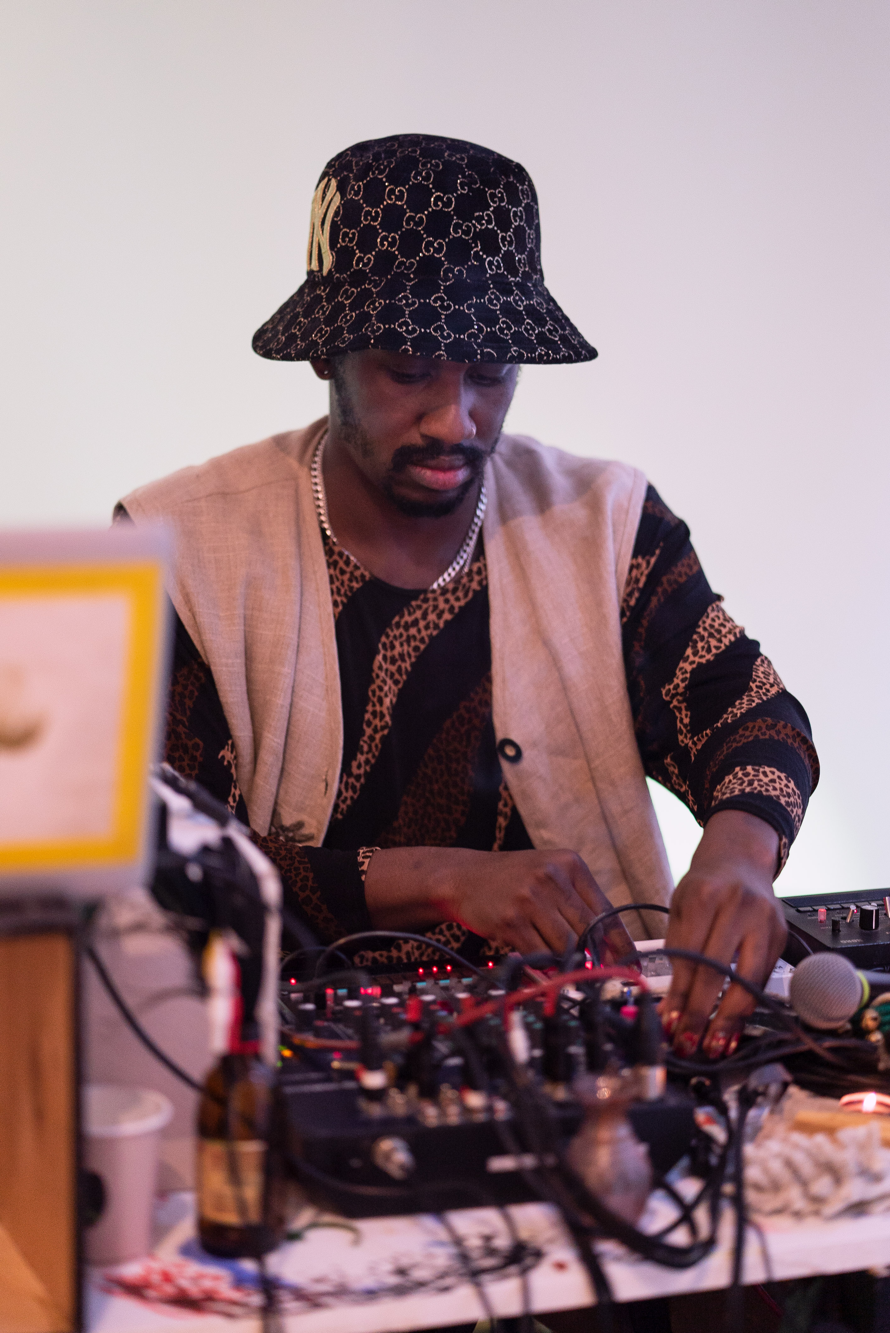 The photo is a medium shot of a person from the front sitting at a DJ table. They are wearing a hat and a traditional vest. In the foreground and out of focus we see equipment, cables and a microphone. The person operates equipment for DJing.
