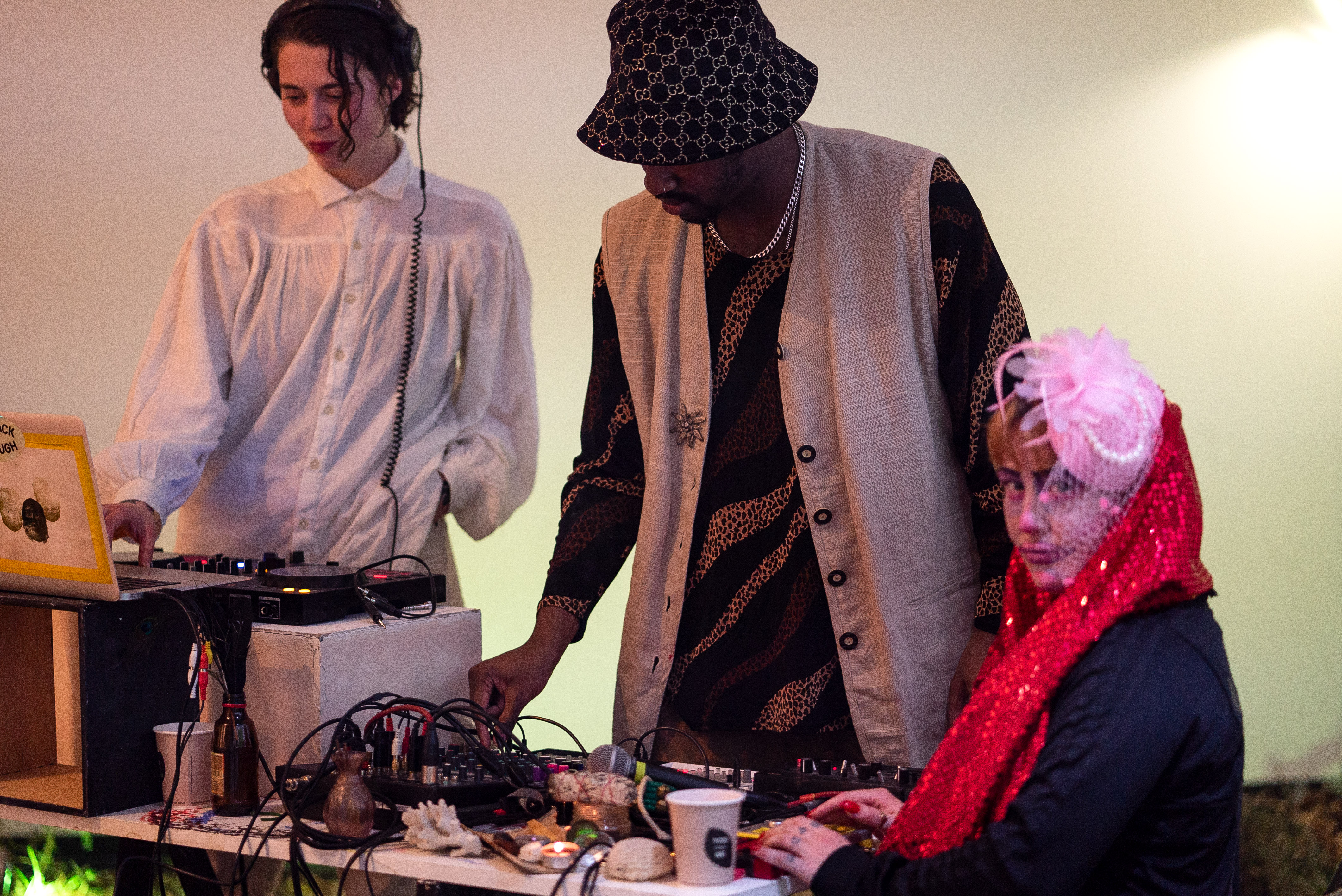 The photo is a medium shot of three people around a DJ table. The person on the far left is wearing white clothes, headphones and has one hand on a laptop. The person in the middle wears a hat, a traditional vest and operates DJ equipment. The person on the far right is the only one sitting at the table and looking towards the camera. She is wearing a large pink flower on her head and has a glittering red scarf wrapped around her neck.