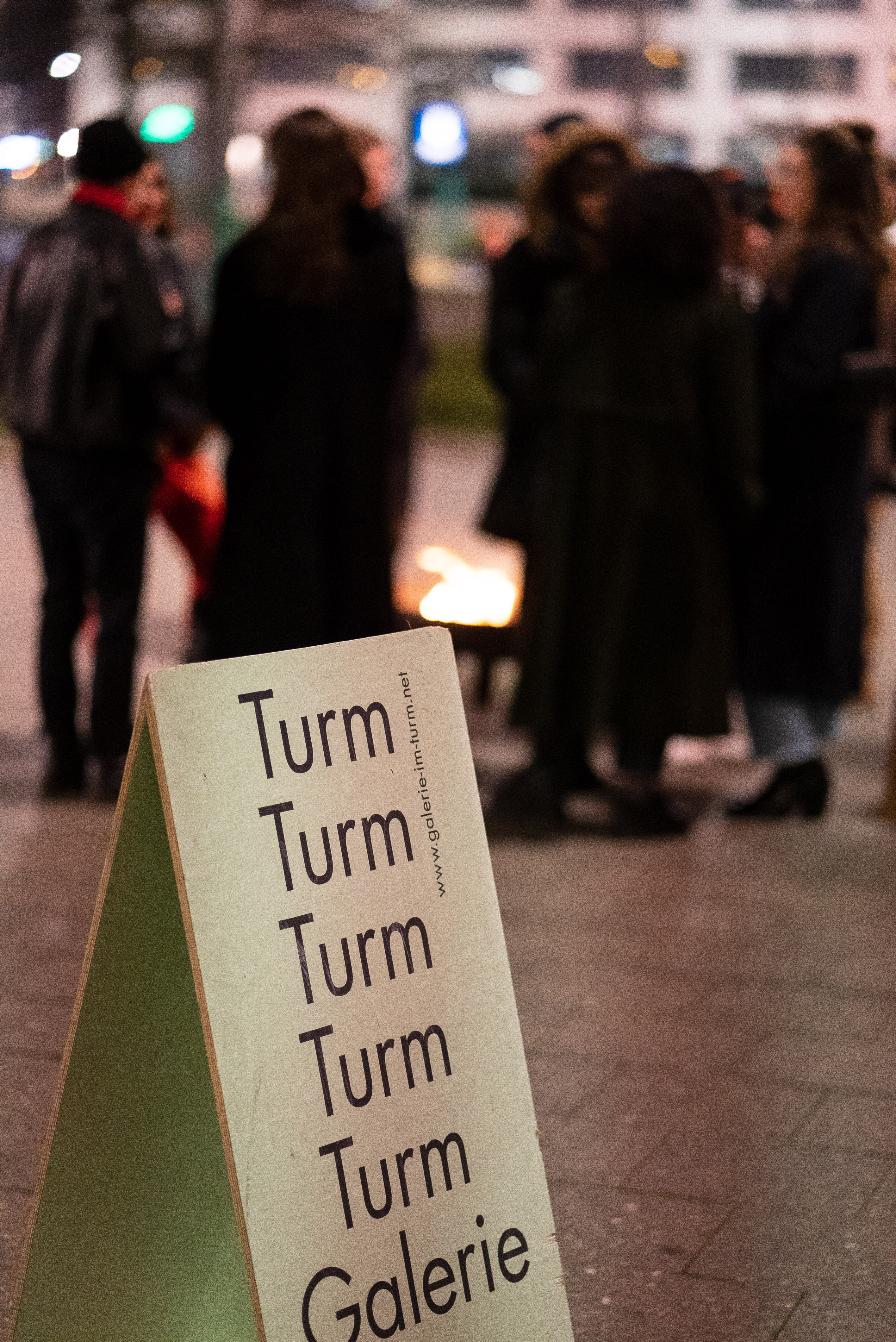 The photo shows in the foreground a sandwich-board sign for the exhibition location, Galerie im Turm. It is evening. In the background in the blur, people stand around a fire bowl.