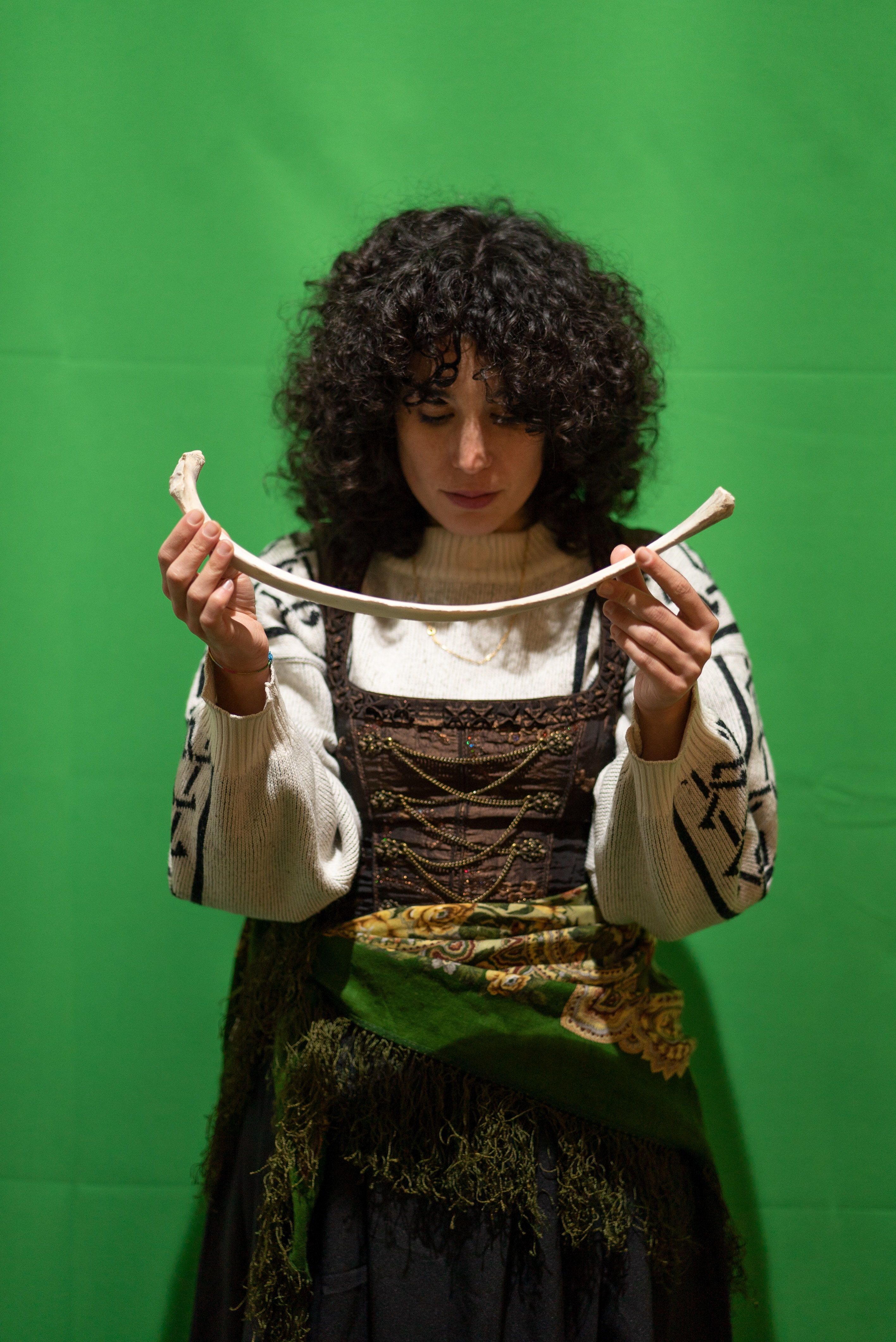 The photo is a medium shot of a person in front of a green screen. They have black curls, wear a wool sweater and over it a traditional costume dress with a scarf around their hips. With their hands they hold a 40cm long bone in front of their face. Their head is lowered and their eyes are directed to the ground.