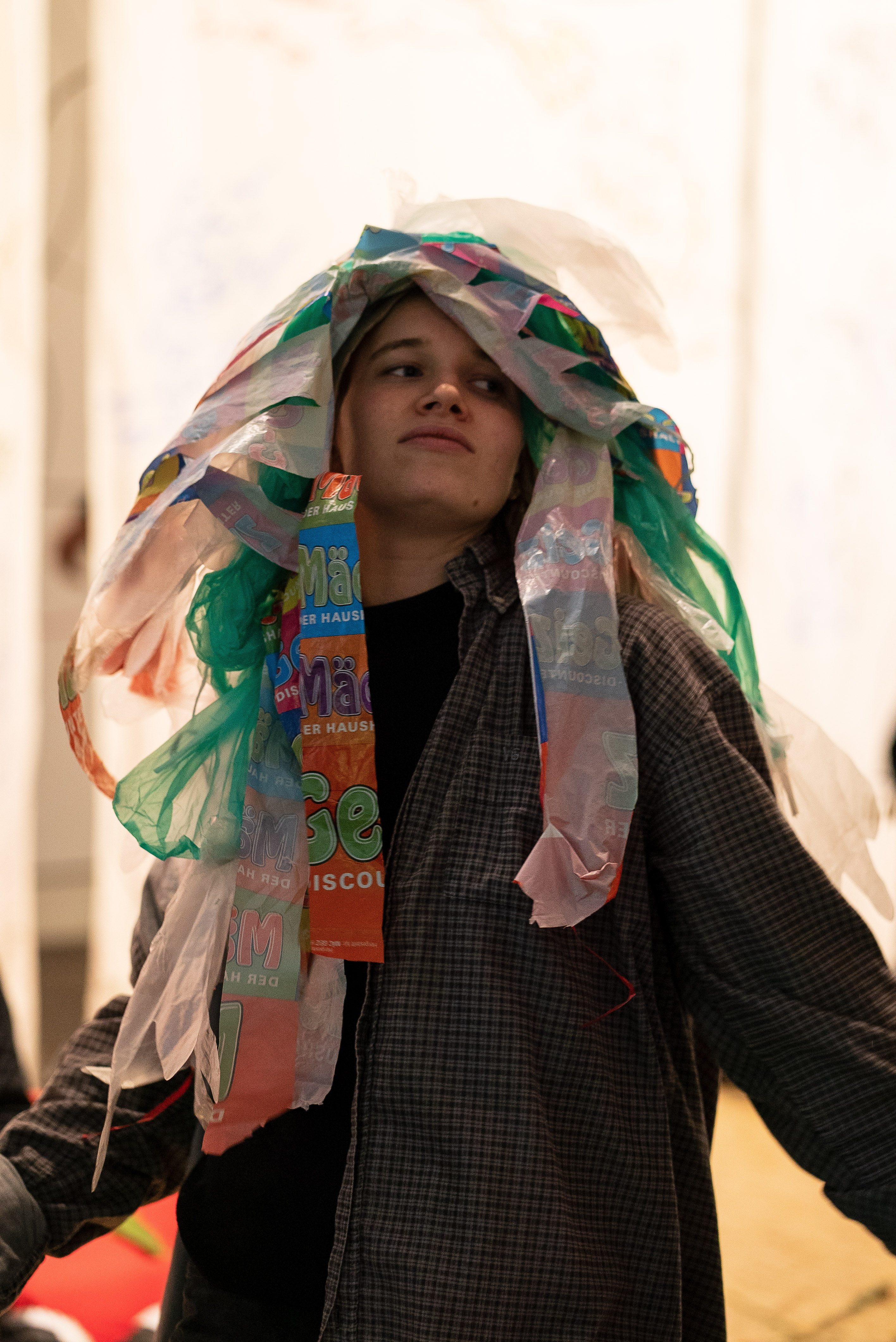 The photo is a medium shot of a person wearing a checked shirt in a dancing position. On the head they wear a colorful headpiece made out of trash bags.