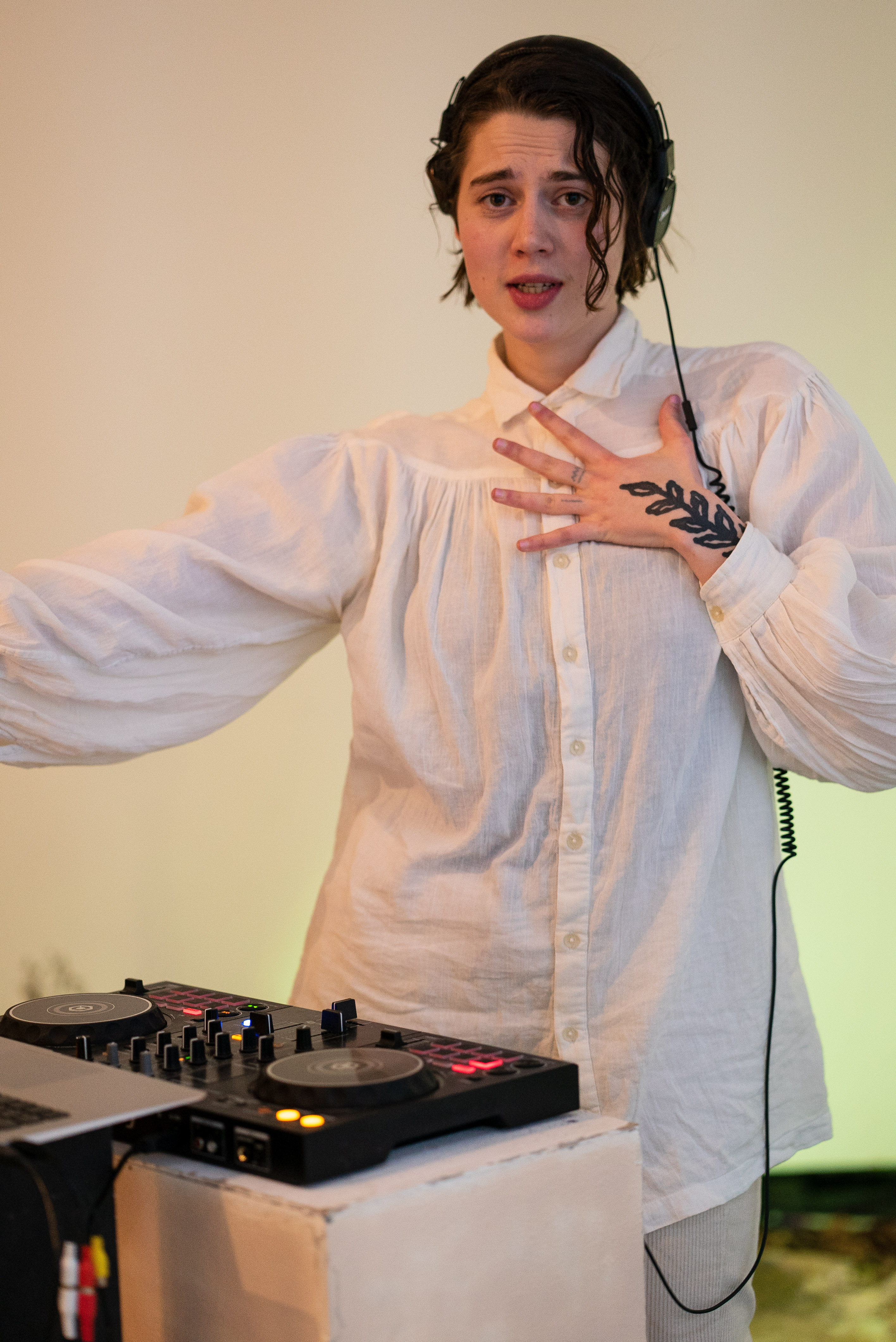 The photo is a medium shot of a person wearing white clothes and headphones dancing in front of a dj table. With the left hand on their chest they are looking into the camera, their mouth looks like as if they were singing.