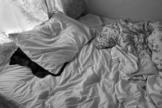 A black and white image of a rumpled bed with white sheets and a floral duvet thrown to the side. The white pillow has another darker pillow peeking out from underneath, and a bright window is behind the bed. The wrinkle pattern in the fabric is most heavily emphasized. A paper with text printed on it is on the far corner of the bed.
