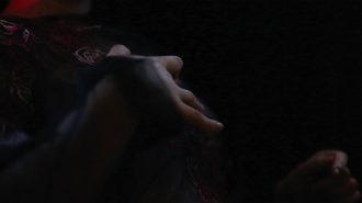 A close up image of the artist performing the piece. In the image, they are pictured using one hand to stroke the wearable suit on their body. A little bit of the pink embroidered detail is visible, but the background is mostly the black suit color. The gauzy fabric of the suit is visible on the arm, and the scene is lit with the light of tv screens. There is slight blur on the hand so that it seems like it is in motion.