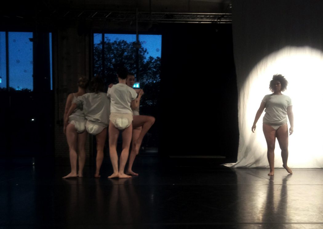 Live performance documentation. In a dimly lit room with a black floor and veiled dark blue windows, four dancers in white t-shirts and diapers stand in a cluster, mostly with their backs to the camera, while another dancer facing forward, is semi illuminated by a spotlight on a white sheet behind them.