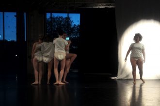 Live performance documentation. In a dimly lit room with a black floor and veiled dark blue windows, four dancers in white t-shirts and diapers stand in a cluster, mostly with their backs to the camera, while another dancer facing forward, is semi illuminated by a spotlight on a white sheet behind them.