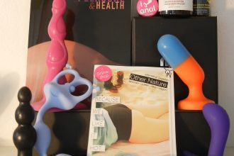 A tableau of books, blocks, anal toys, plugs, beads and lube are arranged against a wall. One book has a view of someone’s bare behind with the title “ANAL PLEASURE & HEALTH” visible. Another title reads “BECAUSE WE LIKE IT HOT” overlaid on a book cover of someone’s thighs, butt, and torso in a top and underwear. Above a block, a pink pin stating “I [graphic of a butt plug] anal” rests against a bottle of lube.