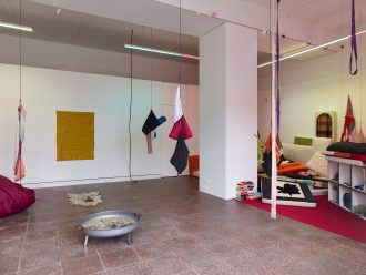 Installation view of a room with varying brightly-colored soft artworks suspended from the ceiling via thin cord or string. In the center, a square column, to the right, a low, freestanding set of shelves with various objects, and piles of pillows. In the foreground, to the left of the column, a freestanding metal fire pit, contents unknown.