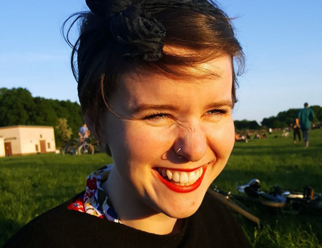 Full-framed portrait of a woman with brown hair and red lipstick standing outside in the sun. She is smiling big and her eyes are squinting. In the background, there is a clear blue sky and folks hanging out recreationally on a grassy field with trees in the distance.