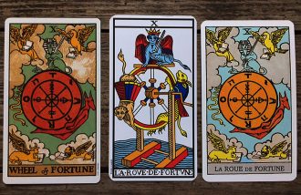 Three Wheel of Fortune tarot cards next to each other on a medium wood surface. The wheel in the first is red with clouds around it, the wheel on the second has dragons on a moving wheel, and the third has orange with a sky in the background.