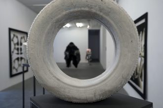 An image of installed art. A cement tire sculpture is visible in the foreground, and other artwork can be seen through the center and around the sides of the sculpture.