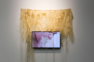 A digital screen with a still from nicola awang's video, depicting a black woman eating blonde hair extensions. On the wall behind the screen, blonde hair extensions are draped.