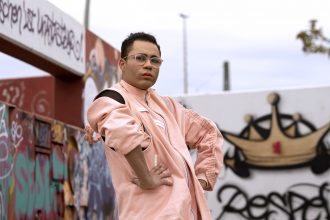 A waist-up picture of a person standing in front of of graffiti'd walls. They have brown skin, dark hair, are wearing glasses, and are looking at the camera, unsmiling. They are wearing a pink outfit with white accents and a black bodysuit underneath.
