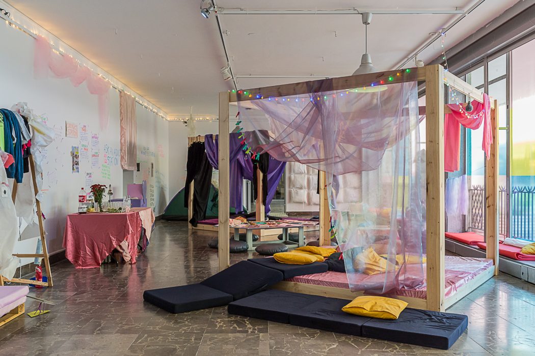 Installation view of Bedtime at Arsenal Gallery. A four poster bed of raw wood is in the middle right of the frame, and sitting pads and pillows spill off the bed into the foreground. There are tables and a view of the gallery walls, with writing and gauze on them. The scene is lit with natural light.