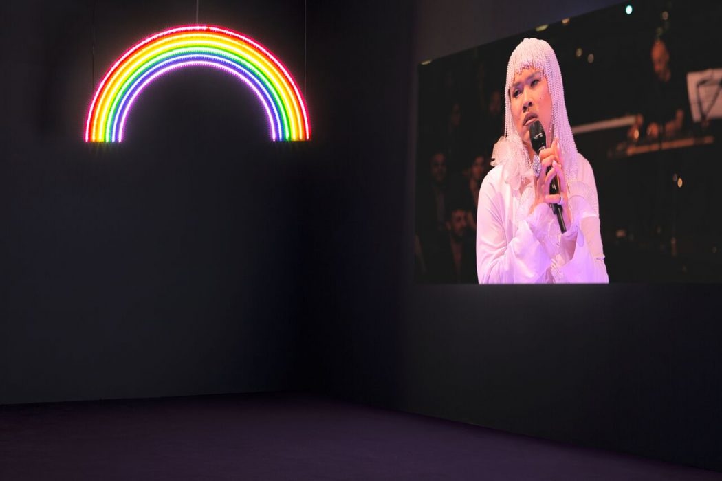 An image of a dark room with grey walls and a purple carpet floor. On the left side of the frame, a glowing LED rainbow hangs down from the ceiling. On the right, a screen with a person in a blonde wig and white outfit using a handheld microphone looks out at the room.