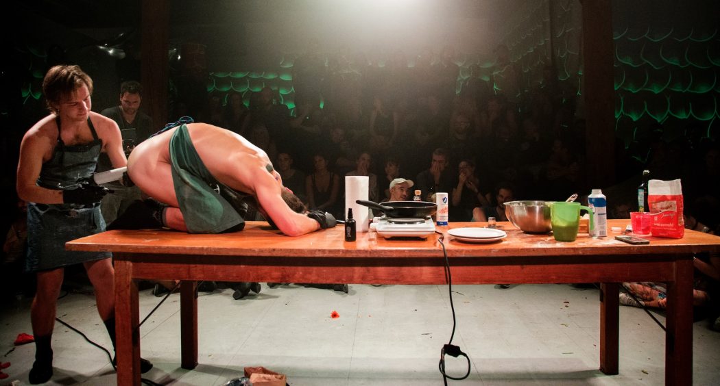 Live performance documentation. The room is dark except for a flood lighting that illuminates a large wooden table, whose left side is strewn with various kitchen implements (hot plate, saute pan, tableware) and baking ingredients and the two nude performers wearing green aprons and black surgical gloves. One person is on top of the left side of the table, knees bent, hunched over with their arms above their head, while the other person is standing behind them on the floor, penetrating their asshole with an implement. In the dim background, three tiers of bleacher style seating show a packed audience of spectators.