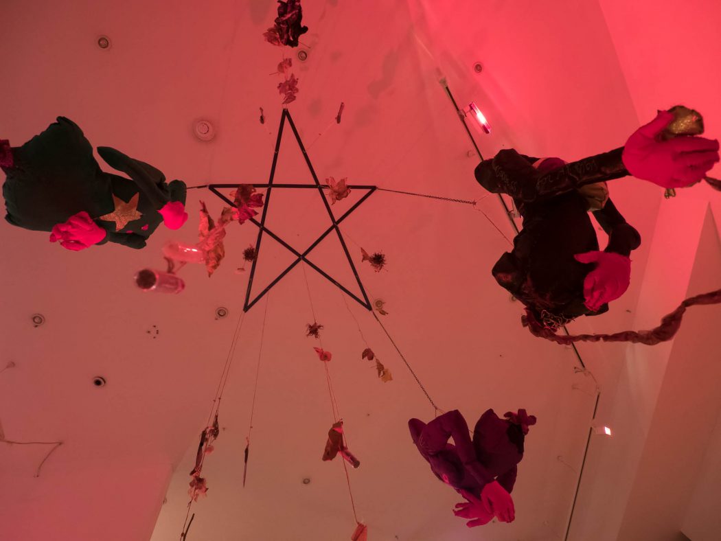 A picture facing up toward the ceiling, which has a pentagram on it. Hanging down from the pentagram are fabric figures, herbs, and other objects.