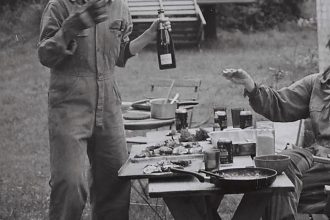 A black and white photograph of two people wearing work jumpsuits. One is standing and holding a bottle of sparkling wine, while the other is reaching for it from the right side of the frame. Neither person's head is visible. The table between is laid with food and dirty plates. They are outside in a grassy, possibly forest area.