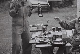 A black and white photograph of two people wearing work jumpsuits. One is standing and holding a bottle of sparkling wine, while the other is reaching for it from the right side of the frame. Neither person's head is visible. The table between is laid with food and dirty plates. They are outside in a grassy, possibly forest area.