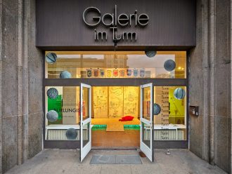 The photo shows an exhibition view of Burlungis from outside. The double doors of the gallery space Galerie im Turm are wide open. Above, the gallery name is lettered in metal on the stone wall. The stage installation by Irene Fernandez Arcas is visible inside.