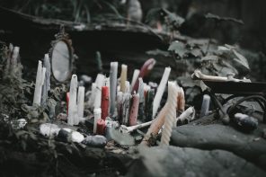 A slightly desaturated close-up image of a rocky outcropping with many candles in reds, pastels, and whites, some textured and some not. Some of the the candles are burnt and wax has spread. Behind them, a small circular mirror in an ornate brass frame is visible, and small flat stones are arranged around the candles. A bone resembling a human femur is visible on the left side of the frame on a ceremonial plate, and natural green foliage is visible in the background.