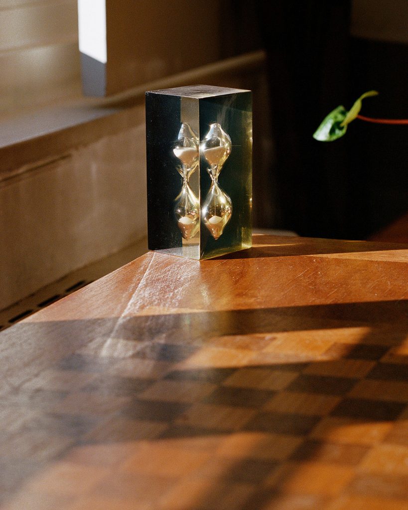 An image of a silvery hourglass-like shape in a translucent, square-shaped material. The object is sitting on a medium colored wooden table in sunlight. A checkered pattern is visible in the shadow on the table, and a plant's leaves are visible on the side of the frame next to the object.