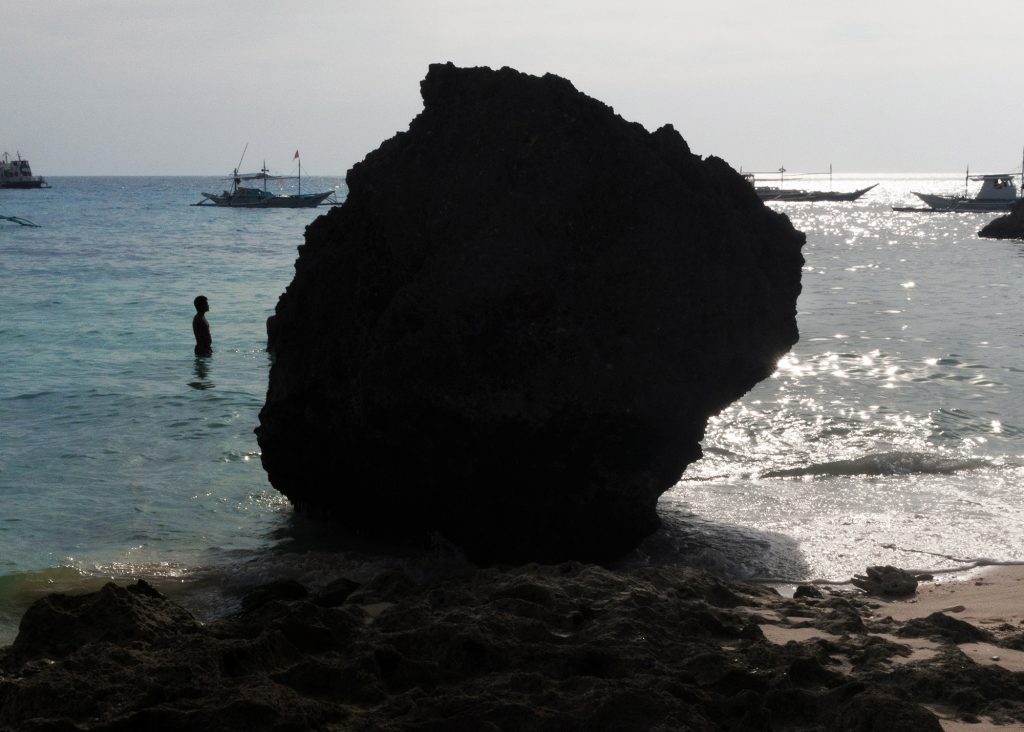 An image of a large, dark boulder sitting in shallow water on a tropical beach. A human and several votes are visible behind the shadowed boulder.