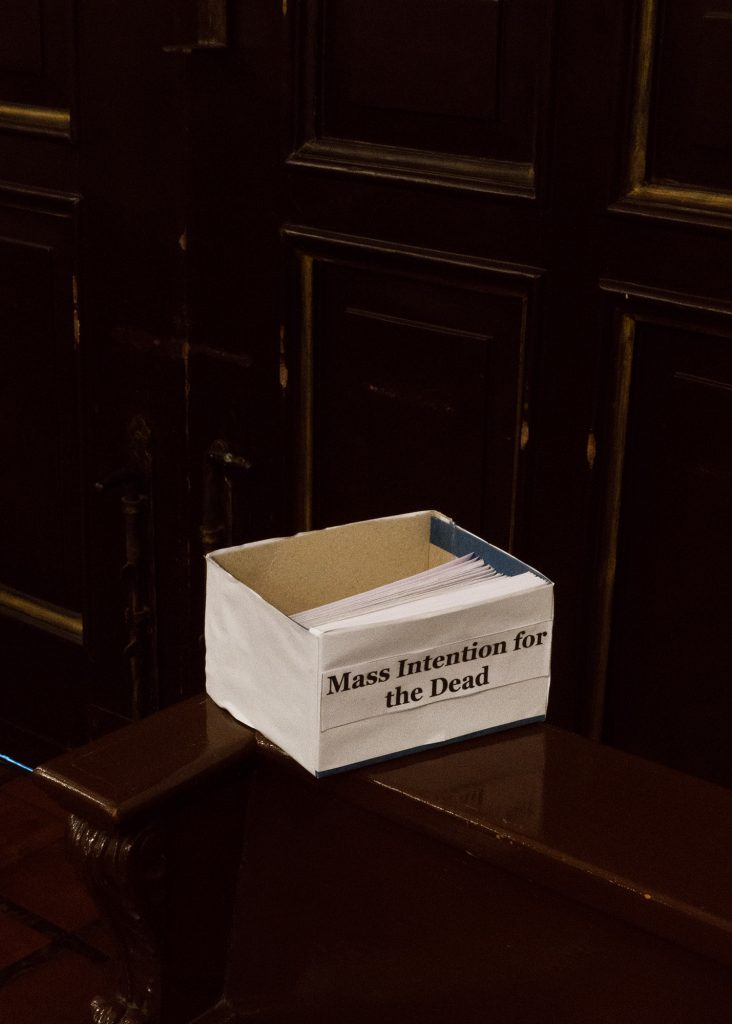 A small paper box on a dark wooden table. The box has a label which reads "Mass intention for the dead". White pieces of paper are stacked neatly inside.