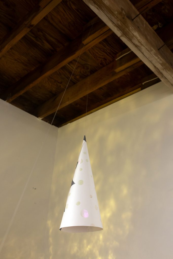 An image of a triangular paper hat suspended from the ceiling, in a room with dappled lighting across the walls. A wooden ceiling is visible above.