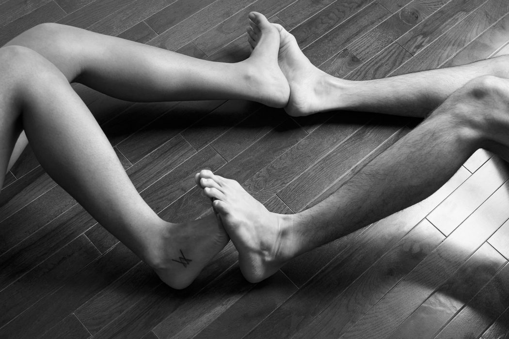 A black and white image of two people pressing their feet together with their lower legs bent to the right and left sides of the frame. They are on a hardwood floor.