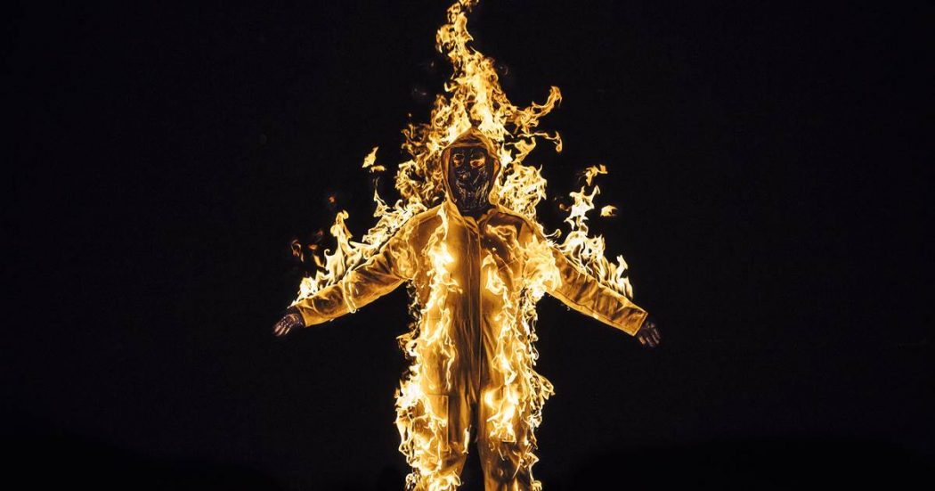 Image from a Cassils of a person on fire againsta a black background, presumably wearing a flame proof suit, arms outstretched.