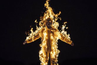 Image from a Cassils of a person on fire againsta a black background, presumably wearing a flame proof suit, arms outstretched.