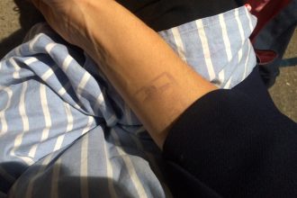 This is a detail shot photo of someone’s wrist, their skin a color like milky coffee, and just barely visible on their inner wrist is a faded stamp, like the kind one may get as they enter a party or club they’ve paid entrance for.