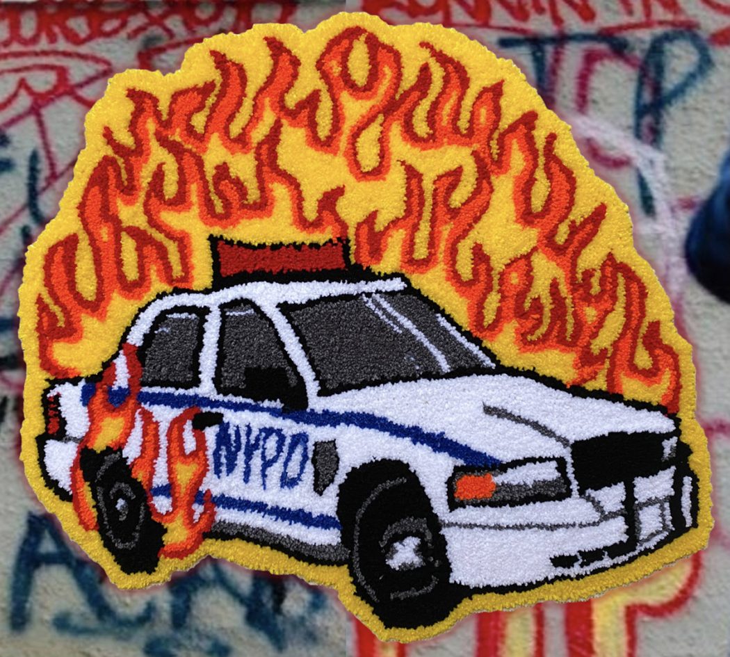 An image of a handmade rug depicting a NYPD cop car on fire with red and yellow flames. The rug is depicted on a graffitied stone wall.