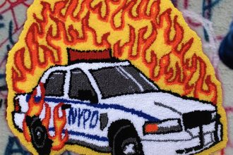 An image of a handmade rug depicting a NYPD cop car on fire with red and yellow flames. The rug is depicted on a graffitied stone wall.