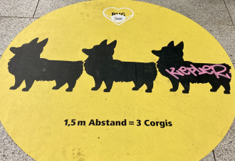 An image of a social distancing messaging sticker in the Berlin subway. The decal features 3 corgis in outline, indicating the appropriate distance.