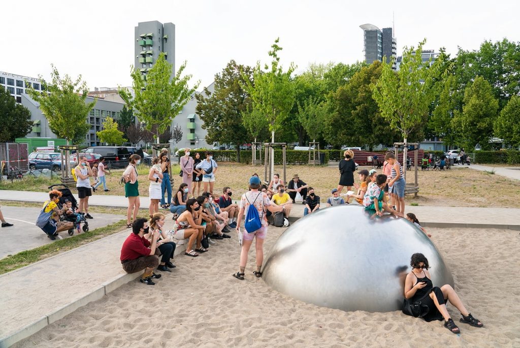 A group watches Frances and Louise performing at a playground. Children are playing on a silver dome behind them.