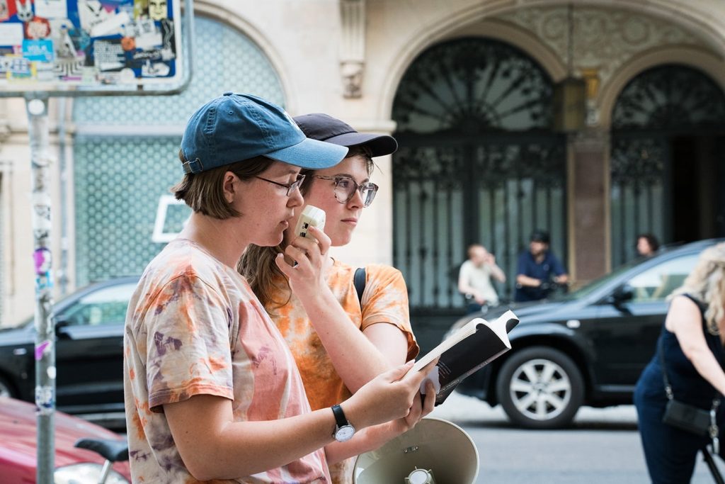 Louise Trueheart, a tomboy femme with long hair in tie-dye, looks at the audience and hold s a microphone up for Frances Breden, a bisexual dyke with medium length hair and also in tye-die, reads a poem.