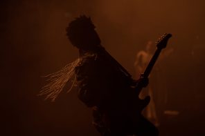 A person in silhouette, bathed in orange light. The image is quite dark, and the person is holding a guitar and gold bead shoulder pads catch the light as they turn toward the camera.