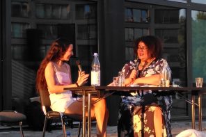 Two people sit at a desk outdoors in front of a gallery, in warm evening summer light. On the left is Anisha, a brownskinned person with long dark hair wearing a pink shirt and skirt and smiling. The other person is Schwarzrund, a Black person with curly black hair, wearing a long black floral dress and talking into a microphone.