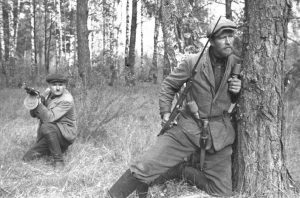 A black and white image of two men holding guns in a forest. One is crouching in the background, while the one in the foreground is leaning against a tree and looking toward the right side of the frame.
