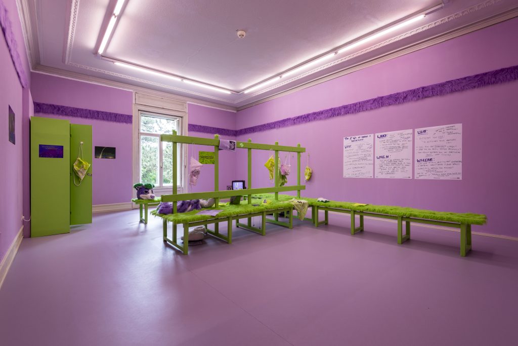 This is a shot of the room from the entrance. We see the benches and lockers, which are the same green color. On the wall are three large pieces of paper that appear to be from a flip chart where hand-written notes on the who, what, when, and where of having a threesome are written, mimicking notes from a coach on a given team’s game strategy.